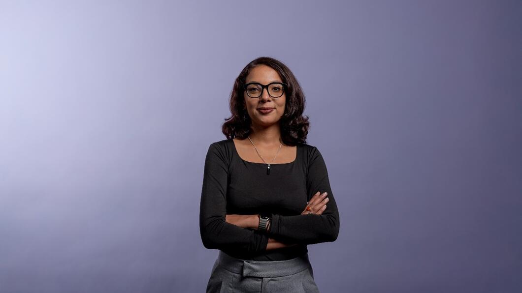 Imane staat met haar armen over elkaar lachend voor een paarse achtergrond