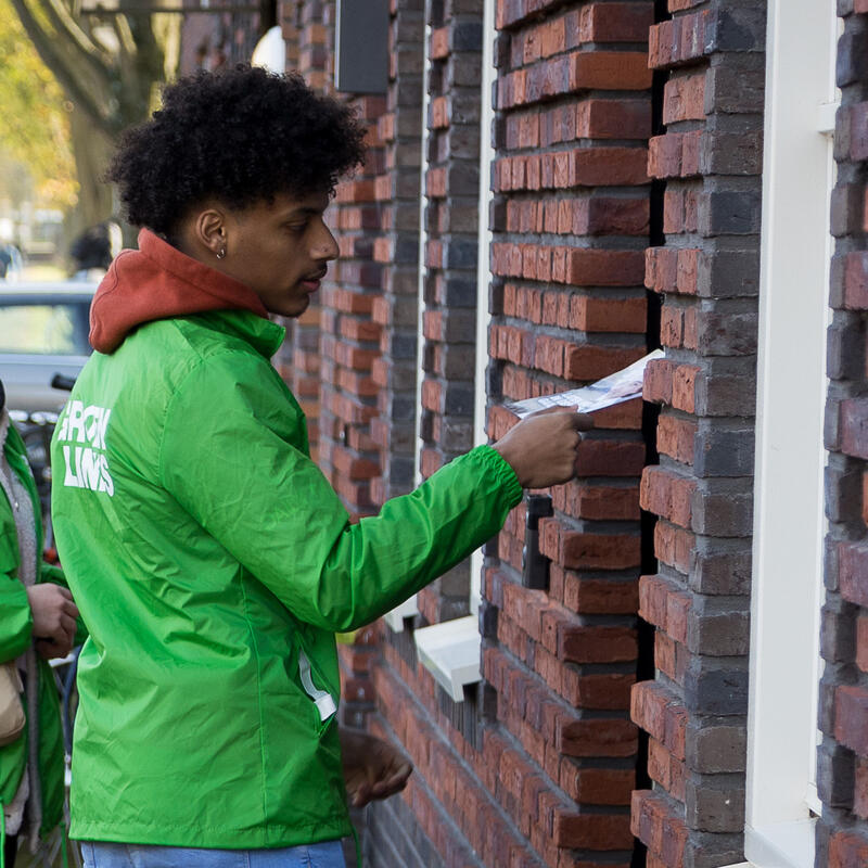 Twee vrijwilligers in een GroenLinks jasje doen een brief in een brievenbus